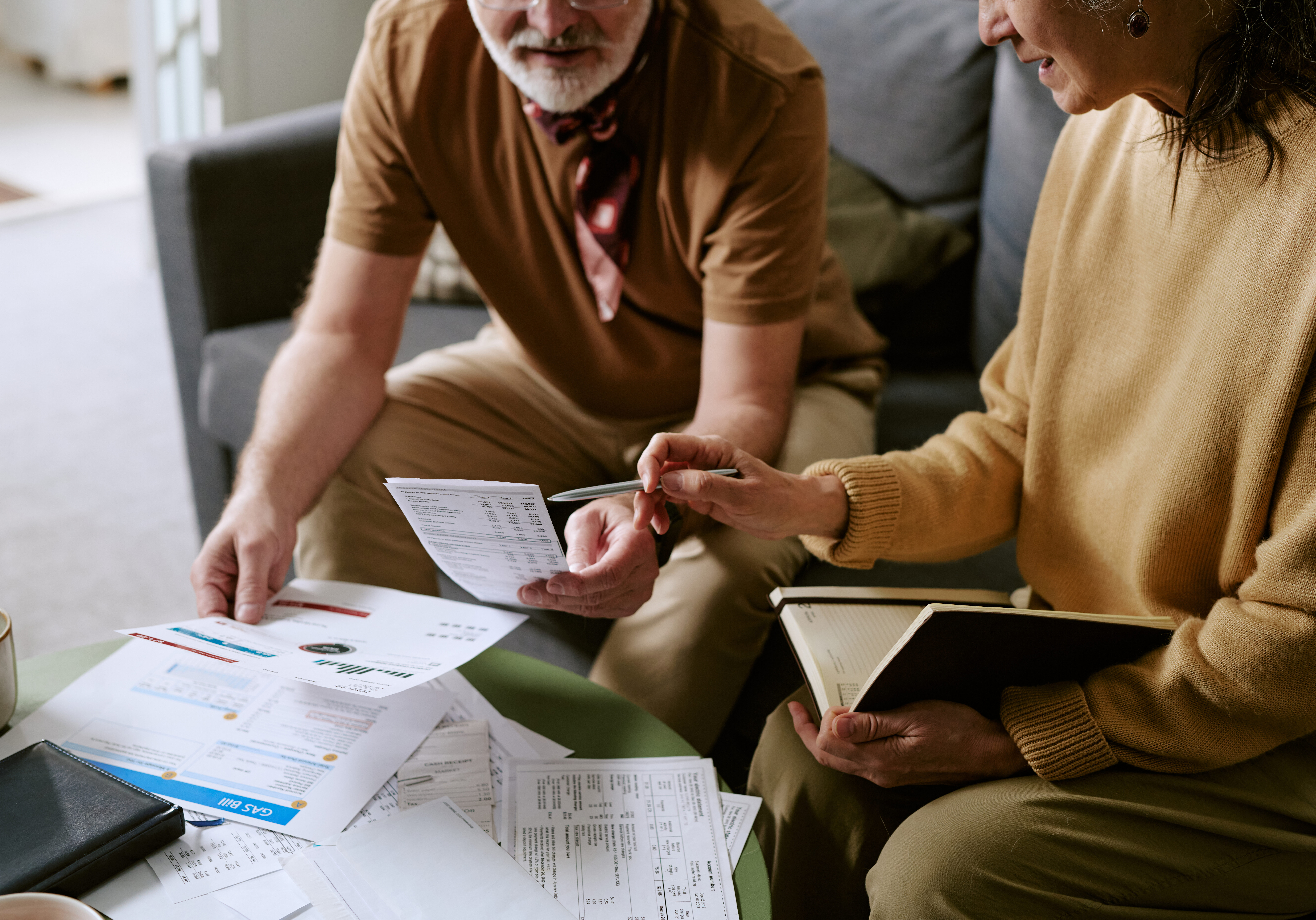 Senior Caucasian man and woman reviewing financial documents together at table, woman holding notebook and pen, man examining printed statements