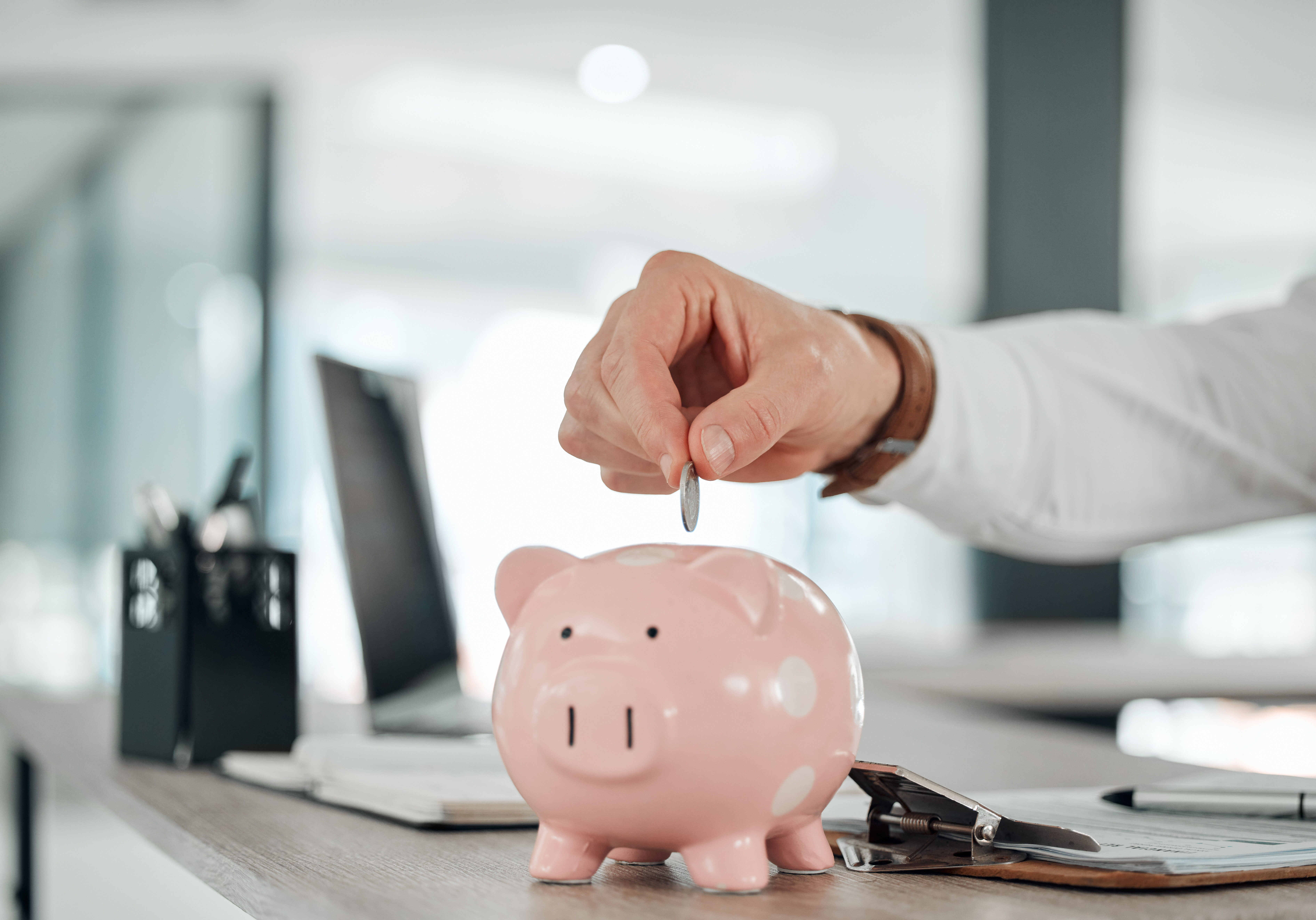 Close-up of a hand dropping a coin into a pink piggy bank on a desk in an office setting